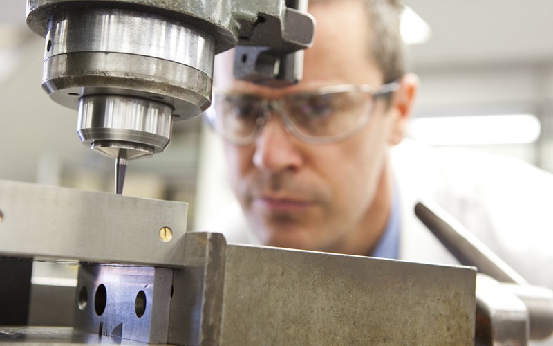 man wearing safety goggles looking at a part being drilled with a pillar drill